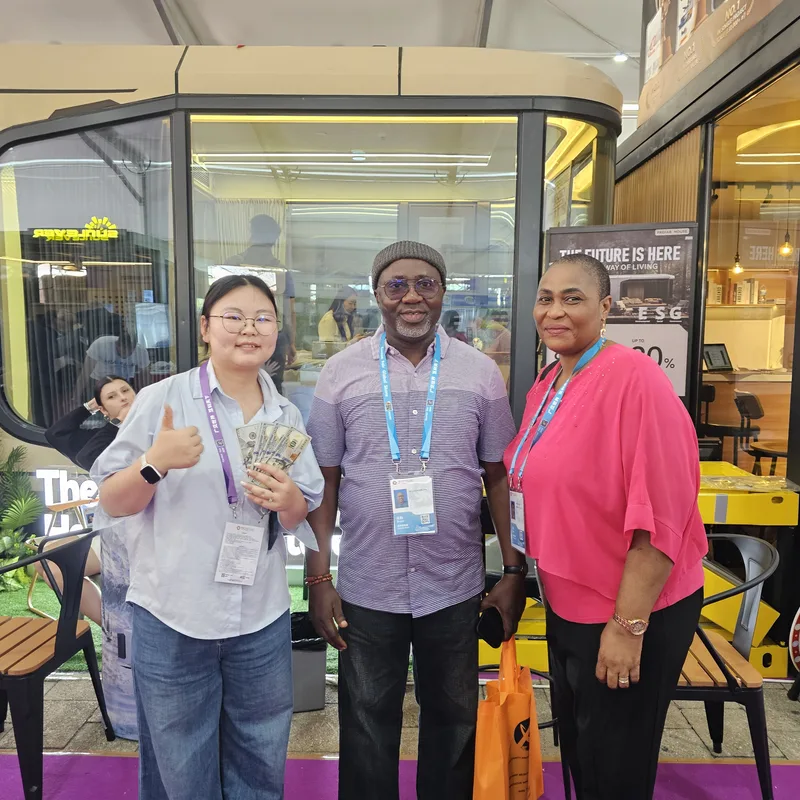CGCH sales rep with thumbs up holding USD cash fan, with African buyer couple at the Z Folding and Detachable container house display